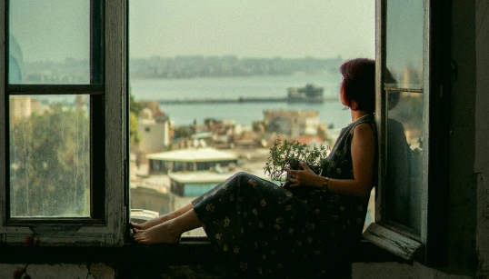mujer-sentada-en-el-marco-de-una-ventana-mirando-la-ciudad-y-el-mar-al-fondo-con-una-maceta-en-las-manos